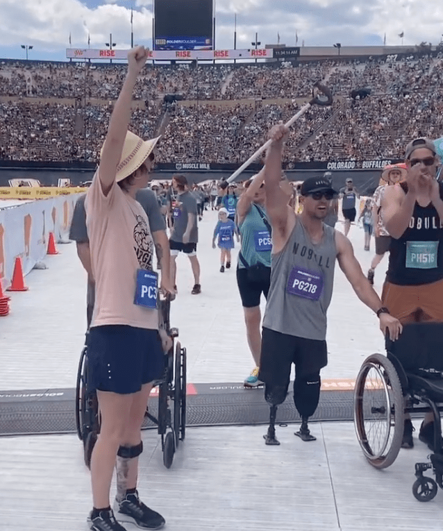 Christa is standing in a straw hat, pink t-shirt and blue running shorts with her fist in the air cheering for Craig, who is on her right in a grey NO BULL tank top and black basketball shorts. Craig is standing on two prosthetic legs, holding onto a wheelchair with his left hand and raising a cane in the air with his right hand. He and Christa have just crossed the finishline of the Bolder Boulder 10k race. There are several race participants finishing behind them.